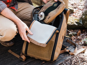 Person opening a coyote brown backpack outdoors with a natural setting