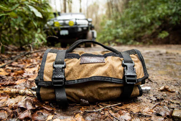 Coyote BROG Tool Pouch Roll secured with buckle straps on a wet, muddy off-road trail, with an overland vehicle in the background.