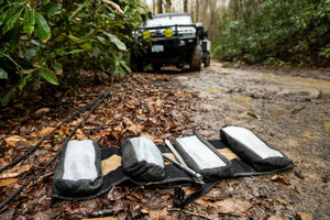 BROG Tool Pouch Roll opened on a muddy off-road trail with four removable tool pouches and hand tools, with an overland vehicle in the background.