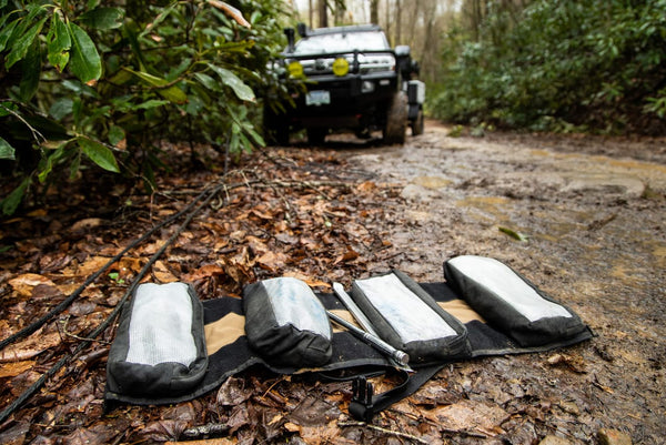BROG Tool Pouch Roll opened on a muddy off-road trail with four removable tool pouches and hand tools, with an overland vehicle in the background.